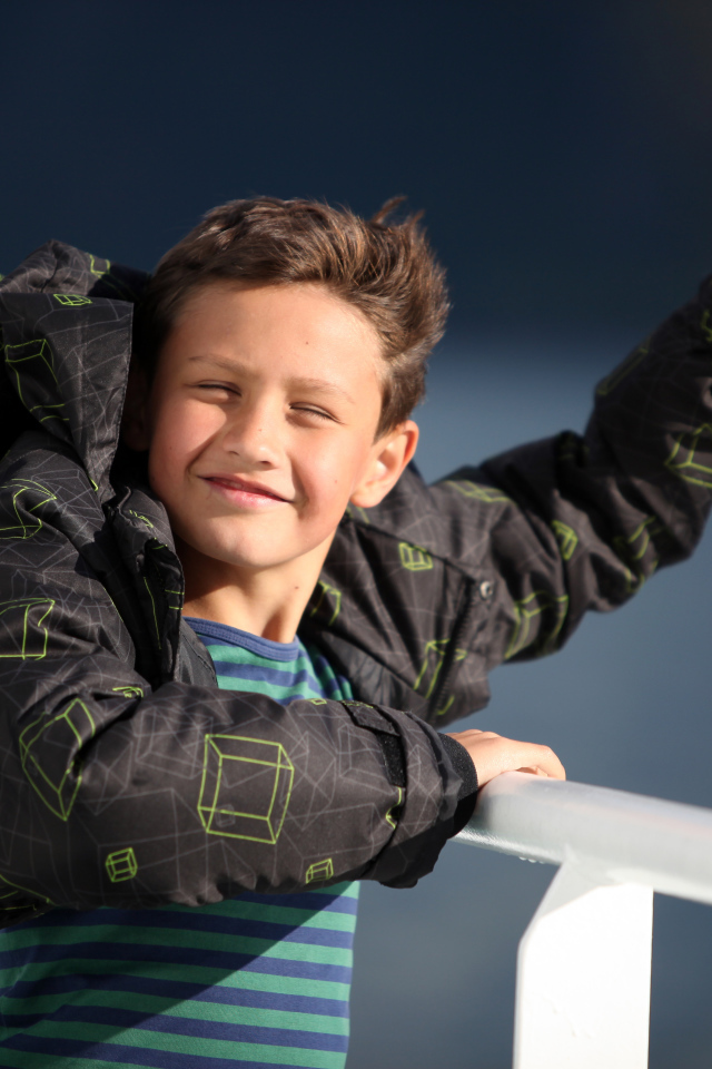 Веселый мальчик в куртке стоит на ветру в лучах солнца