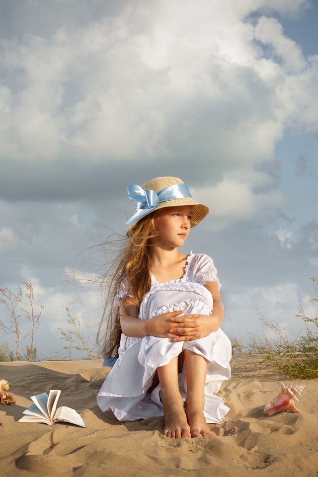 Маленькая девочка в белом платье сидит на песке 