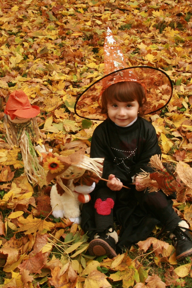 Маленькая девочка в костюме ведьмы сидит на опавшей листве на Хэллоуин