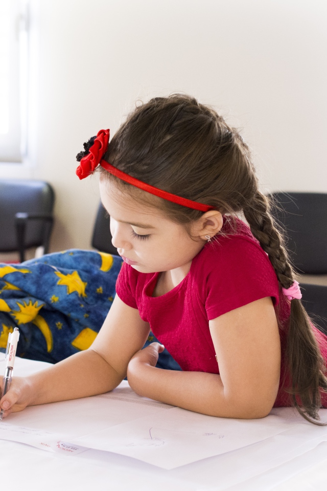 Маленькая девочка в красном платье рисует за столом