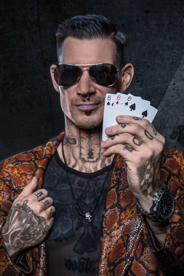 Мужчина с татуировками на теле держит в руке три карты шестерки 
