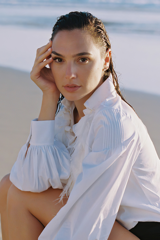 Актриса Галь Гадот в белой рубашке сидит на песке