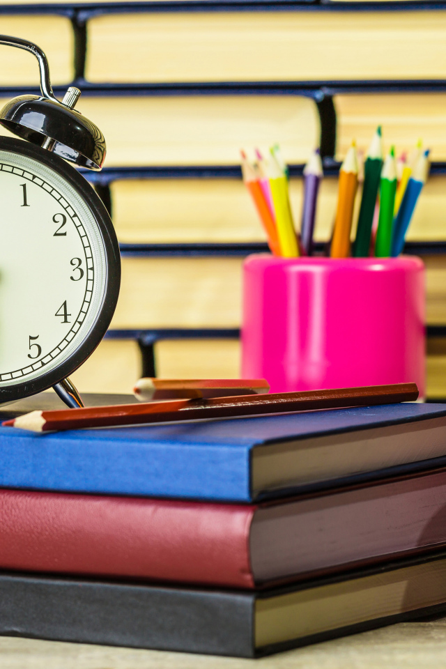 Будильник, книги, карандаши и яблоко на столе у стены 