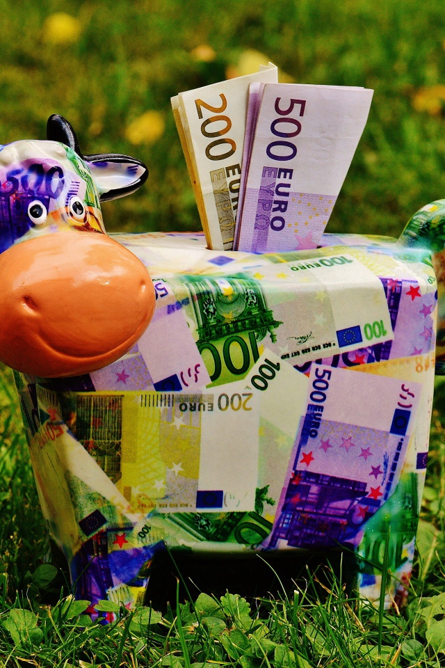 Копилка корова с купюрами евро на зеленой траве 