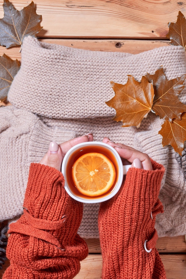 Чашка горячего чая с лимоном в руках на столе с теплым свитером