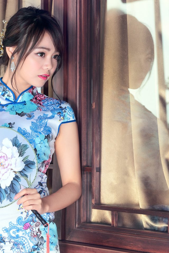 Девушка азиатка в красивом платье стоит у окна 
