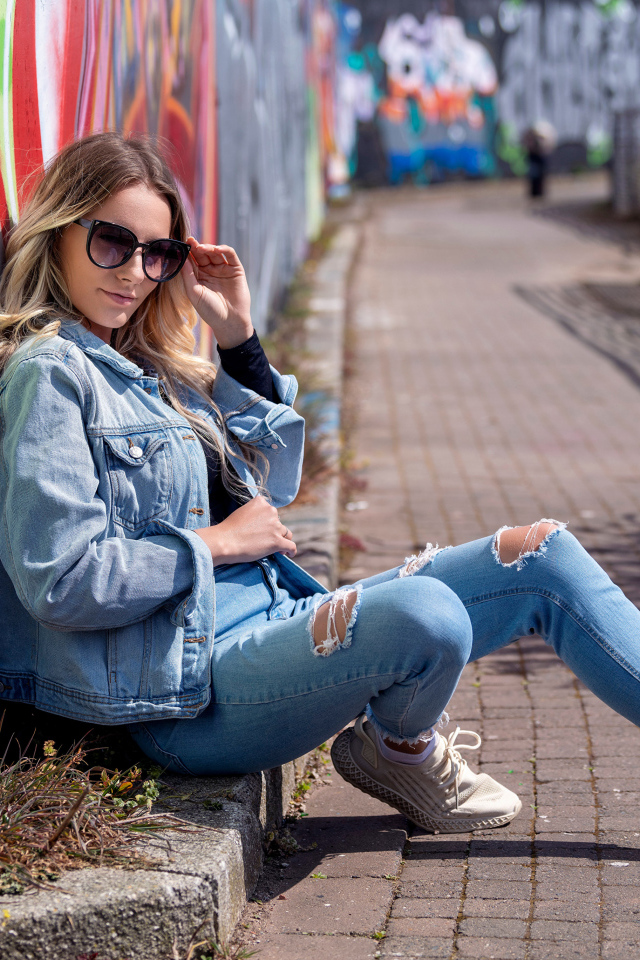 Красивая девушка в джинсовом костюме сидит у стены 