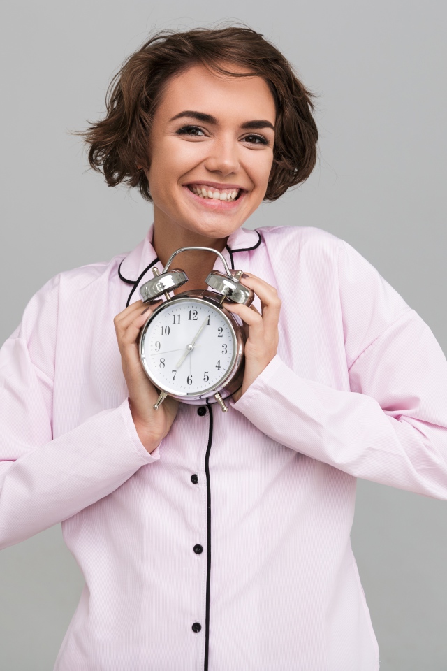 Улыбающаяся девушка в розовой рубашке с будильником в руках 