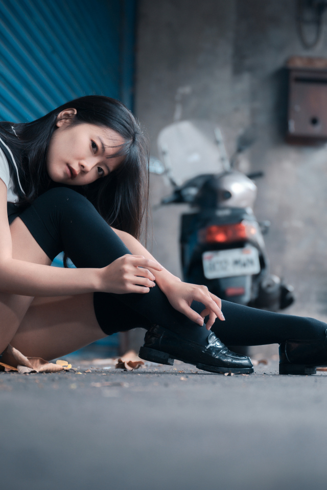 Молодая азиатка в черных чулках сидит у стены 