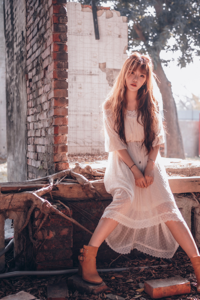 Молодая девушка азиатка в белом платье сидит на разваленной стене