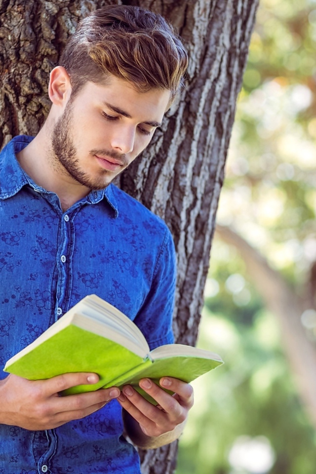 Красивый молодой мужчина стоит с книгой под деревом 