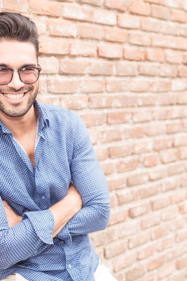 Улыбающийся мужчина в очках стоит у стены 