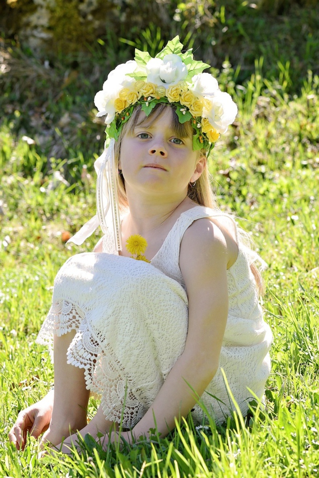 Маленькая девочка с венком на голове сидит на зеленой траве