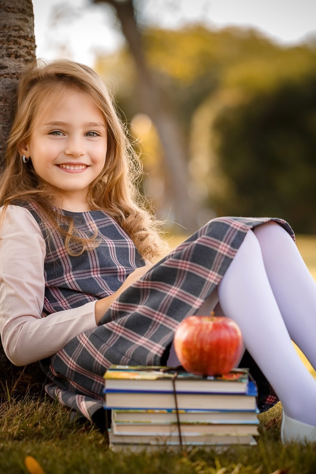 Маленькая девочка с книгами и яблоком сидит под деревом 