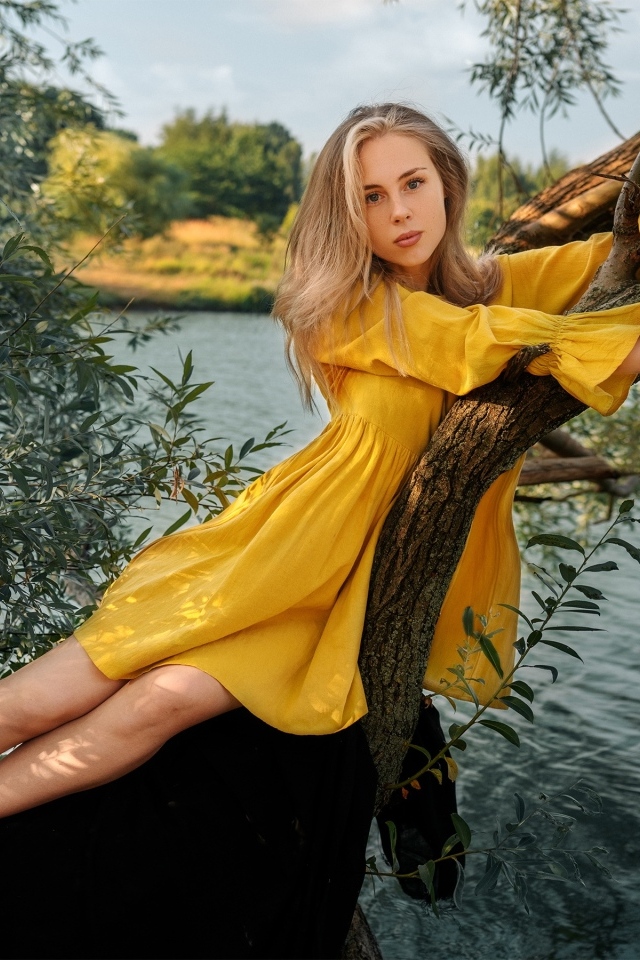 Красивая девушка в желтом платье лежит на дереве у воды