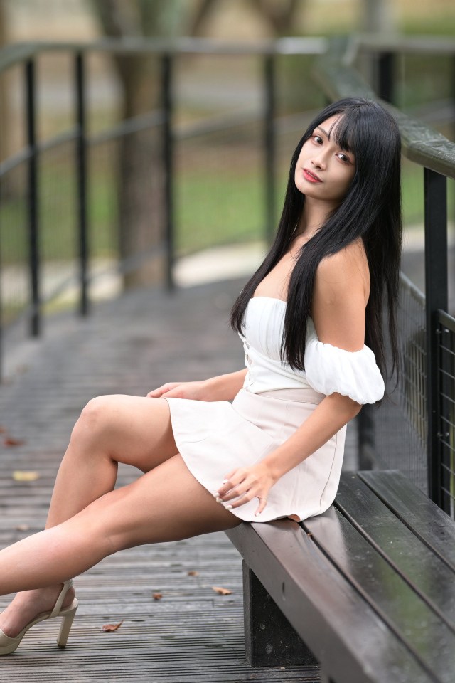 Красивая длинноволосая азиатка в белом платье сидит на лавке