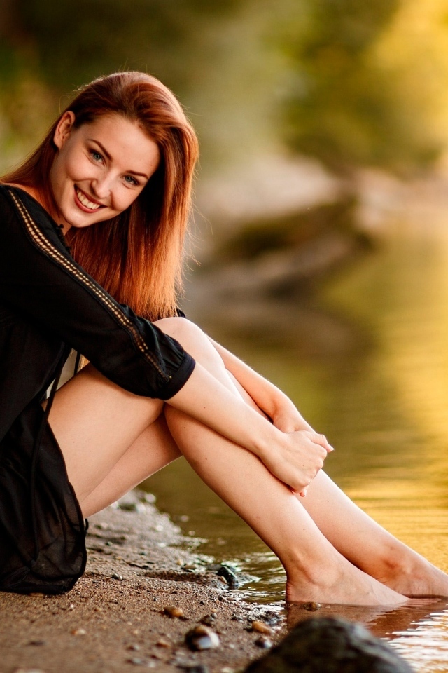 Улыбающаяся девушка в черном платье сидит у воды