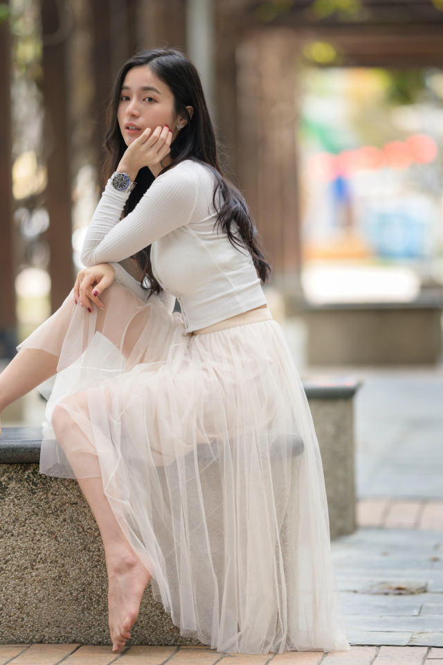 Девушка азиатка в белой юбке из фатина сидит на лавке