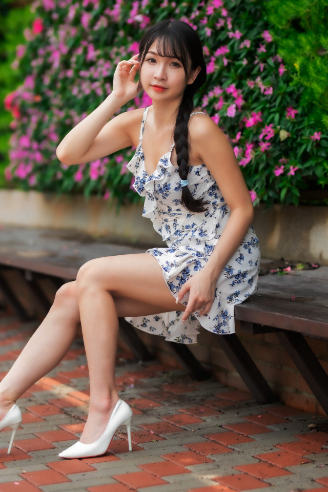Азиатка в платье сидит на лавке у цветущих растений