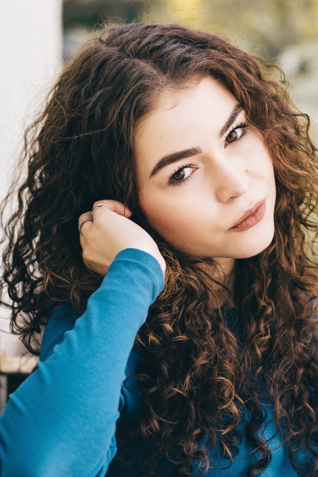 Кареглазая девушка с кудрявыми волосами стоит у стены