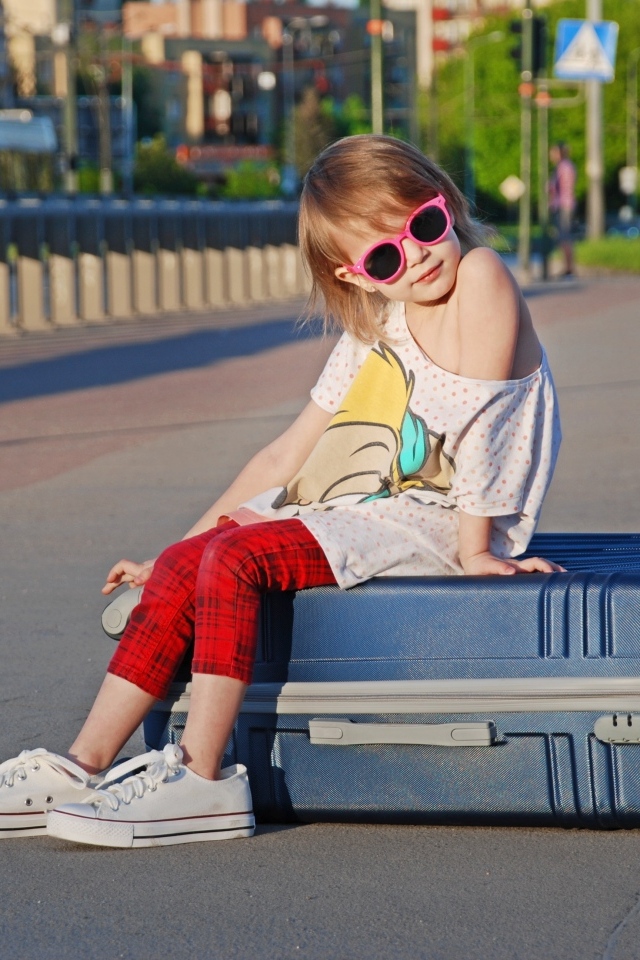 Девочка в солнечных очках сидит на чемодане на дороге
