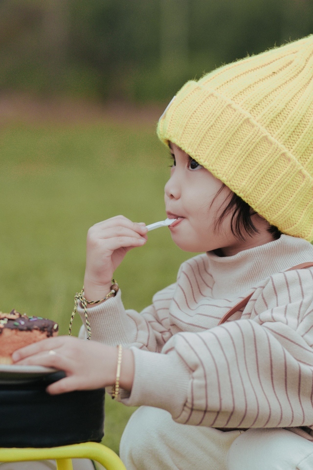 Маленькая девочка в желтой шапке ест пирожное