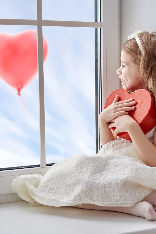 Маленькая девочка с коробкой в форме сердца сидит у окна