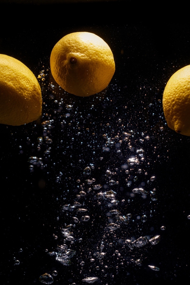 Три желтых лимона с пузырями в воде на черном фоне