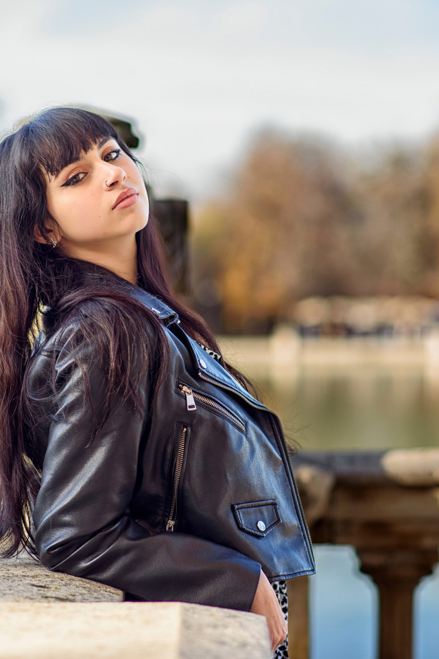 Девушка в черной куртке стоит на мосту у воды