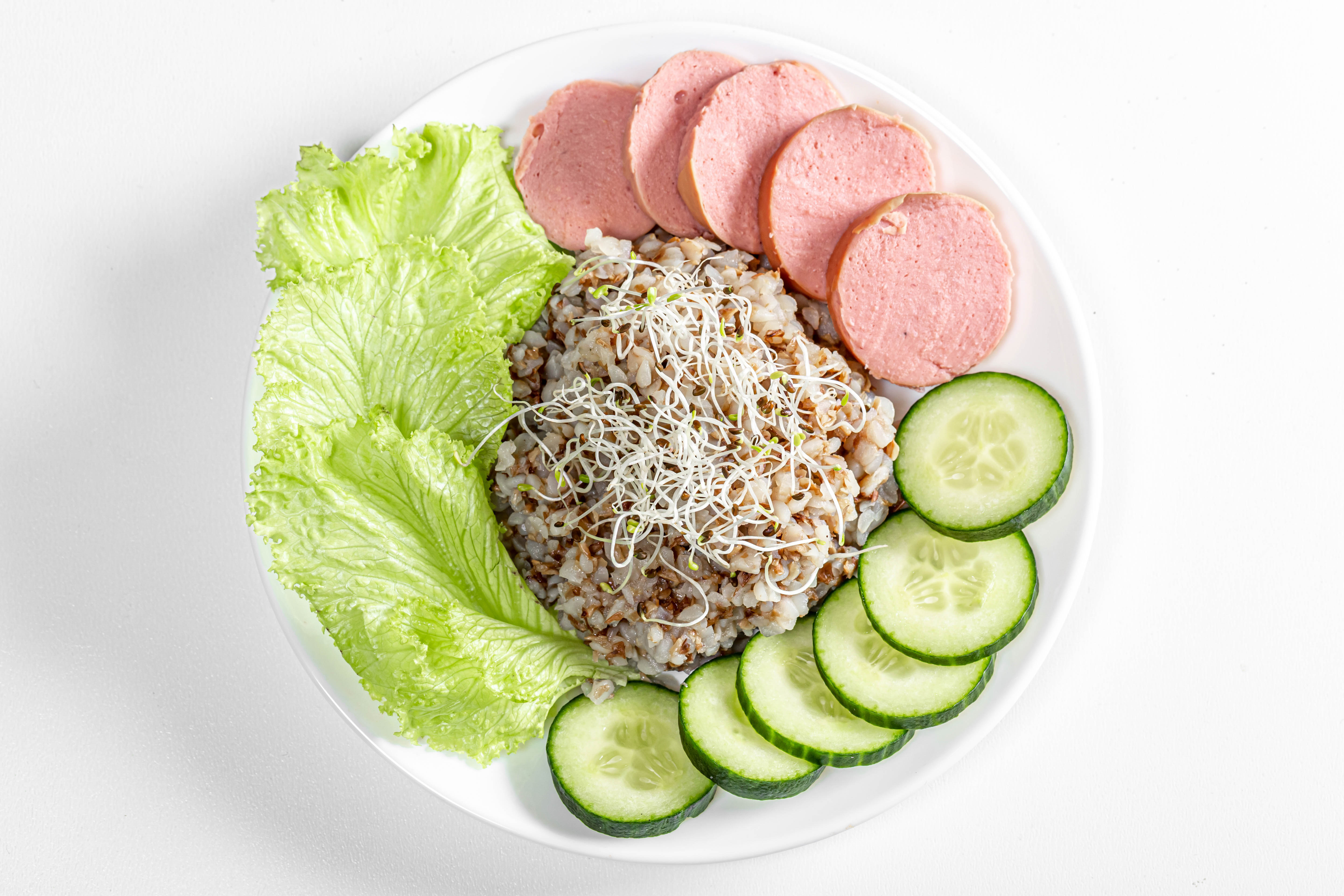 Zastaki.com - Гречка на тарелке с огурцом, колбасой и листьями салата на белом фоне